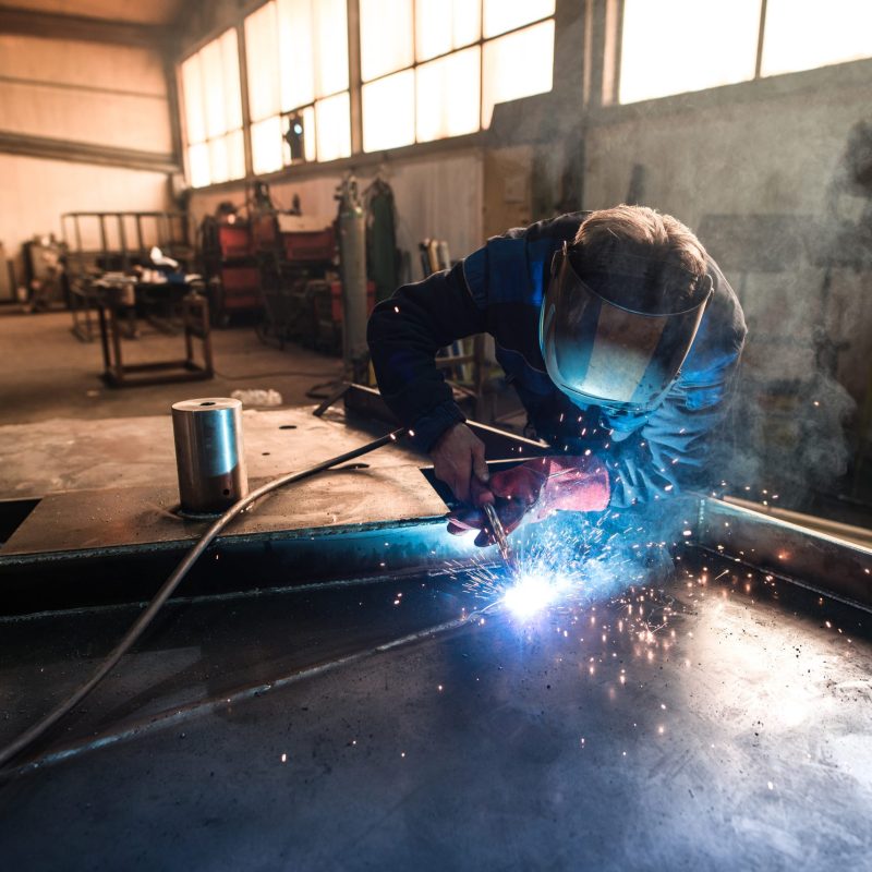 Professional welder welding metal construction parts in industrial workshop.